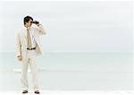 Businessman using binoculars, standing on beach