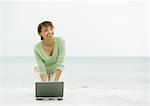 Woman on beach, kneeling on sand with laptop
