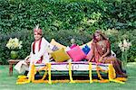 Portrait of a newlywed couple in traditional wedding dress sitting on the bed in a lawn