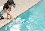 Little girl sitting by edge of swimming pool, touching surface of water