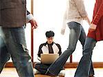 People Passing by Man Using Laptop on Floor