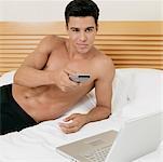 Young man lying on the bed using a remote control with a laptop in front of him