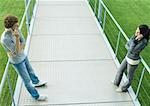 Teenage couple standing face to face on walkway, using cell phones, full length, high angle view