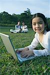 Girl lying in grass using laptop, parents sitting in grass in background