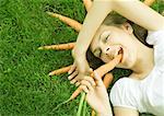 Woman lying on grass, eating carrot, carrots arranged around head, smiling at camera