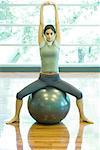 Young woman sitting on fitness ball, stretching arms overhead