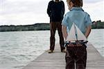 Little boy is standing in front of a man with a miniature sailing boat behind his back, both on a footbridge, selective focus
