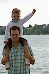 Mid adult man is standing in front of a lake with a boy on his shoulders, selective focus