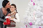 Mother kissing daughter's forehead next to a white Christmas tree