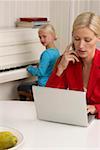 Blond girl is playing piano behind her mother who is working with a laptop, selective focus