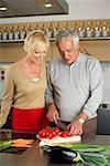 Gray-haired man is cutting tomatoes in a kitchen while standing next to a woman