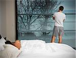 Man Looking Out of Window While Woman Sleeps in Bed