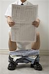 Mid section view of a young man sitting on the toilet seat reading a newspaper