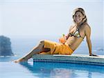 Portrait d'une jeune femme assise au bord de la piscine et tenant un verre de jus