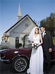 Portrait of a newlywed couple standing near a car in front of a chapel