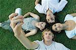 Group of young friends lying on grass, heads together, one young man taking photo with cell phone