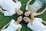 Group of young friends lying on grass with heads together, eyes closed, one taking photo with cell phone