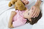 Girl lying in bed with teddy bear, man holding her hand on putting other hand on her forehead