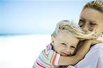 Woman holding daughter on beach, girl smiling at camera as wind tousles her hair