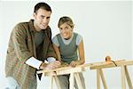 Couple leaning on sawhorses, using measuring tape, smiling at camera
