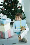 Boy sitting next to stack of Christmas presents, holding one against chest