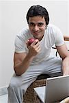 Portrait of a young man holding an apple and sitting in front of a laptop