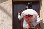 Rear view of a man hiding a bouquet of flowers behind his back and knocking the door