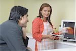 Young woman putting a cake into the oven with a young man looking at her