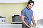 Portrait of a young man standing in the kitchen and using a laptop