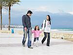 Mother and father walking along palm lined pavement by the sea with young daughter (6-8). Alicante, Spain.