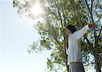Jeune homme debout à l'extérieur, bras tendus, les arbres et le soleil en arrière-plan