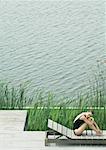Woman sitting on lounge chair next to lake, hugging knees, smiling