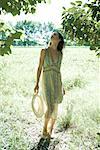 Young woman wearing sundress and holding sun hat, standing looking up at trees