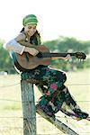 Young hippie woman sitting on fence post, playing guitar