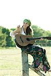 Young hippie woman sitting on fence post, playing guitar