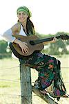 Young hippie woman sitting on fence post, playing guitar