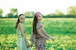 Young hippie women walking through field, holding hands, looking over shoulders at camera