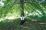 Businessman sitting under tree, laZSop on lap, stretching arms