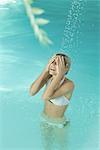 Young woman in pool, standing under stream of water, covering eyes