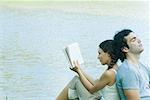Séance de couple dos à dos à côté de l'eau, femme lisant