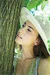 Young woman wearing sun hat, touching face to tree, close-up
