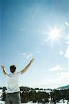 Young man standing in snowy landscape, arms raised to sun, rear view