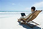 Businessman sitting in lounge chair on beach, barefoot, using laZSop