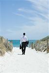 Unternehmer, die zu Fuß in Richtung Strand, holding von Aktenkoffer und Mantel über Arm, Rückansicht