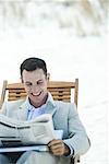 Businessman sitting in lounge chair, reading newspaper, smiling