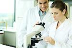 Weibliche Lab Arbeiter Platzierung Folie unter Mikroskop, männlichen Kollegen über die Schulter schauen lächelnd