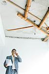 Man in suit standing under bare pipes in home interior, using phone and holding document