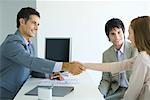 Businessman shaking hands with young woman across desk, young woman's male companion smiling