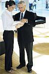 Mature businessman and young businesswoman standing, looking at file in office lobby, full length