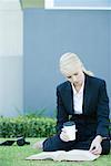 Businesswoman sitting on grass reading book, holding coffee cup